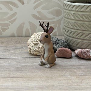 May include: A brown and white figurine of a hare with small antlers, seated on a wooden surface. The hare has large black eyes and is positioned in front of a decorative rock and a textured pot.