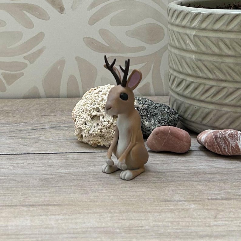 May include: A brown and white figurine of a hare with small antlers, seated on a wooden surface. The hare has large black eyes and is positioned in front of a decorative rock and a textured pot.