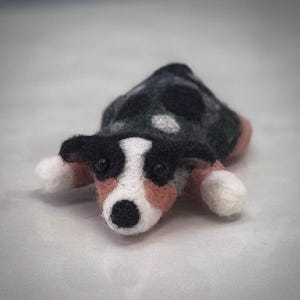 May include: A handmade felted dog figurine in a lying down pose. The dog has a black and white coat with pink accents on the ears and paws. The dog's eyes are black, and the nose is black. The figurine is made of wool.
