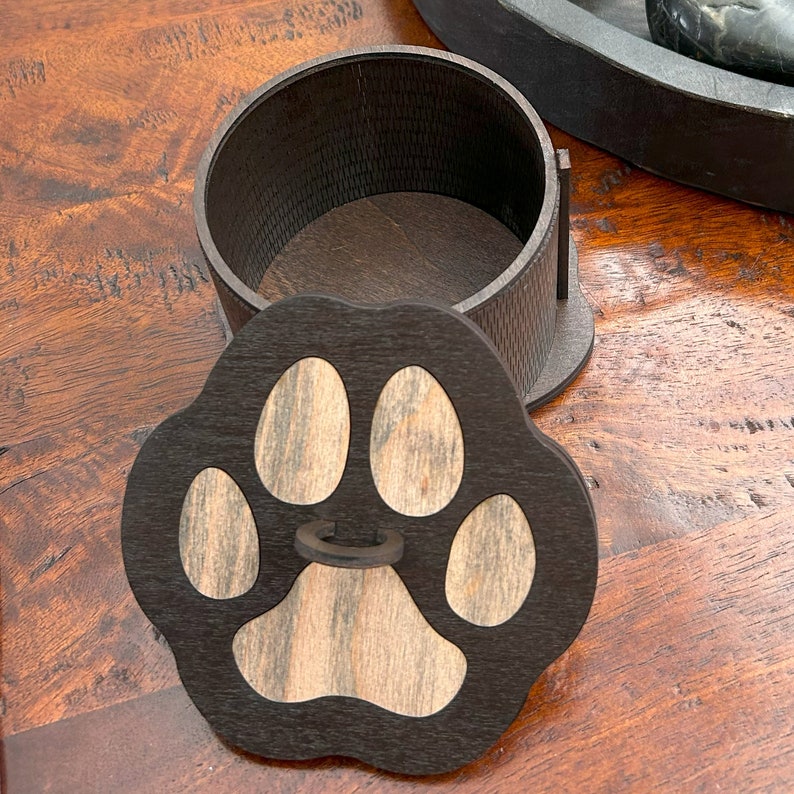May include: A wooden box with a paw print lid. The box is made of dark wood with a light wood inlay. The paw print is black with a light wood center.