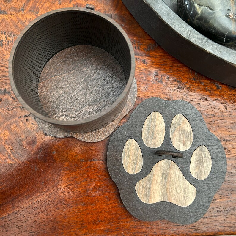 May include: A wooden box with a paw print lid. The box is dark brown with a light brown wood grain pattern. The paw print is made of light brown wood and is set against a dark brown background.