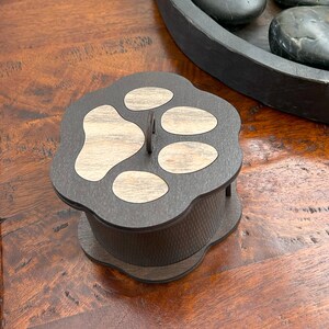 May include: A dark brown wooden box shaped like a paw print with a light brown wood inlay. The box has a small metal loop on the top.