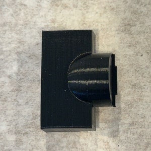 Puede incluir: Un conector de plástico negro impreso en 3D con un extremo redondeado.