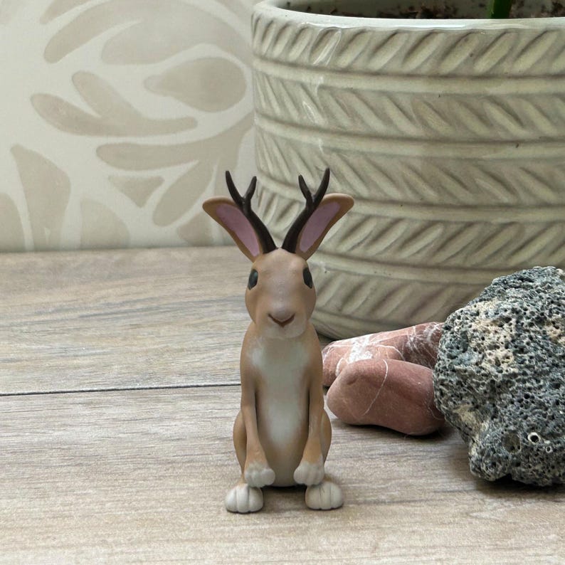 May include: A decorative jackalope figurine, a mythical creature with rabbit features and antlers. The jackalope is light brown with white details and dark brown antlers. The figurine is on a wooden surface.