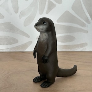 May include: A detailed figurine of an otter standing upright. The otter is primarily dark brown with a lighter brown chest and face. Its paws and tail are also dark brown. The figurine is on a wooden surface.