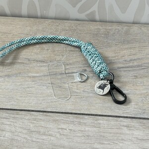 May include: A green and white patterned lanyard with a silver key ring and a silver charm with a black silhouette of a wolf's head. The lanyard is on a wooden surface.