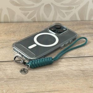 May include: A clear phone case with a black phone inside. The phone case has a white ring on the back for a magnetic phone holder. A teal blue lanyard is attached to the phone case.