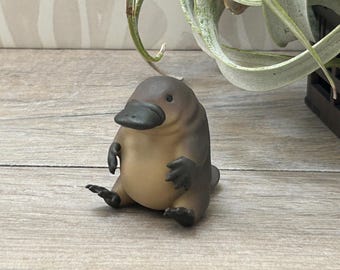 May include: A small, detailed figurine of a platypus in a seated position. The figurine has a brown body, a tan belly, and a dark grey bill and feet. The platypus is sitting on a wooden surface.
