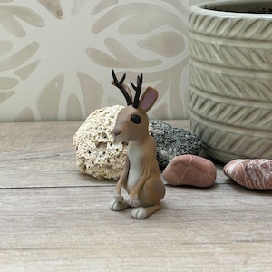 May include: A miniature brown jackalope figurine, a mythical rabbit-like creature with antlers, sits upright. The figurine has black eyes and is set against a backdrop of decorative stones and a textured pot. The jackalope is a whimsical decorative item.
