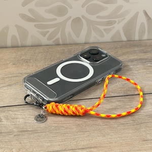 May include: A clear phone case with a black phone inside. The case has a white ring on the back for a magnetic phone holder. A yellow and red braided phone strap is attached to the case.