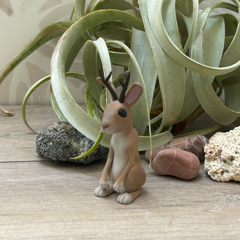 May include: A small, tan-coloured figurine of a rabbit with deer antlers. The rabbit is sitting upright with its ears perked up. The figurine is on a wooden surface, with a green plant and rocks in the background.