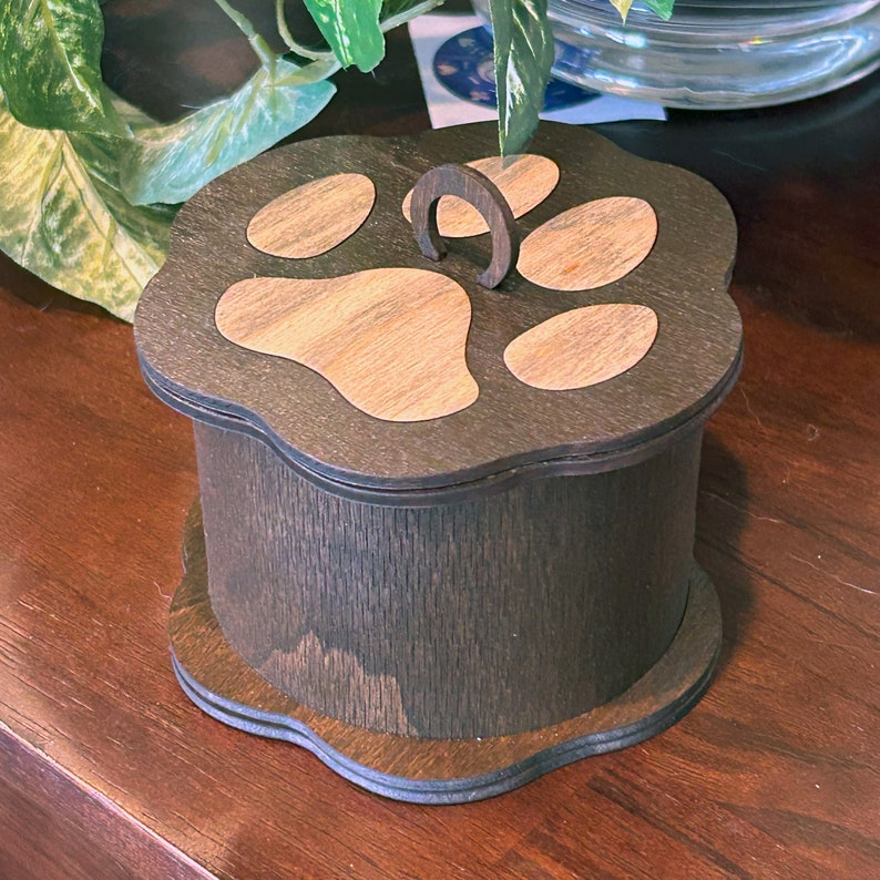 May include: A dark brown wooden box with a paw print design on the lid. The box has a small metal loop on the top.