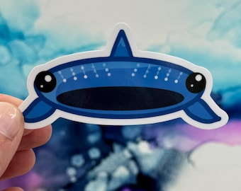 May include: A blue sticker featuring a cartoon-style whale or fish with large black eyes and a white outline. The design includes a dark blue mouth and a dorsal fin. Small white dots are arranged along the top of the whale.