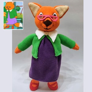 May include: A plush toy fox wearing a green cardigan, a white collared shirt, and a purple dress. The fox has pink glasses and red shoes.