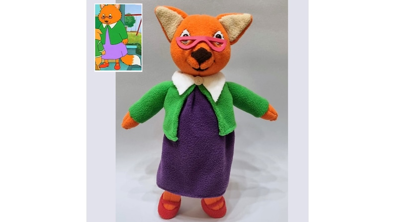 May include: A plush toy fox wearing a green cardigan, a white collared shirt, and a purple dress. The fox has pink glasses and red shoes.