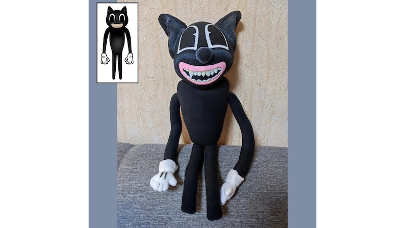 May include: A black cartoon cat plush toy with a wide, toothy grin. The toy has white eyes and a pink tongue. The toy is sitting on a gray surface.