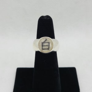 May include: A silver ring with a white face featuring a black Japanese character. The ring is displayed on a black velvet ring holder against a white background. The Japanese character is a stylized symbol.