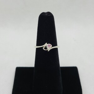 May include: A silver ring with a heart-shaped design. The ring features a small open heart and a cluster of pink gemstones. The ring is displayed on a black ring holder against a white background.