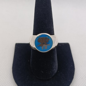 May include: Silver ring with a round, blue enamel face featuring a brown Japanese character. The ring is displayed on a black velvet ring holder against a white background.