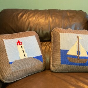 May include: Two square throw pillows with nautical designs. One features a lighthouse with a red top, the other a sailboat. Both have a blue, white, and brown color scheme and a brown border.