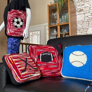 May include: Four knitted throw pillows with different sports themes. The pillows are red, blue, and white with black accents. The sports featured are soccer, football, baseball, and hockey.