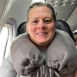 May include: A gray knitted travel neck pillow with a drawstring closure is around a person's neck. The pillow is designed to provide support and comfort during travel. The person is sitting in an airplane seat.