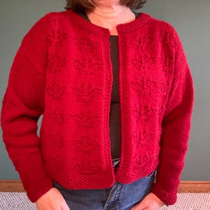 May include: A bright red knit cardigan featuring a textured pattern of raised floral-like motifs across the front panels. The garment has long sleeves, a round neckline, and a straight open front design with a ribbed border along the edges.