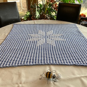May include: A blue and white gingham throw blanket with a white snowflake design in the center. The blanket is lying on a white table with a small toy bee in the foreground.