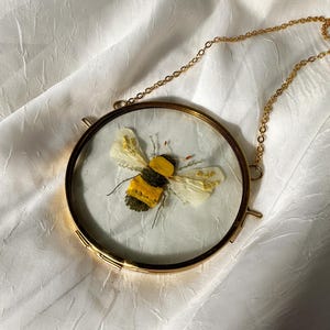 May include: A circular wall hanging featuring a pressed bee made of yellow and green dried botanical elements. The bee is encased between two clear glass panes within a thin gold-toned metal frame, suspended by a matching gold-toned metal chain.