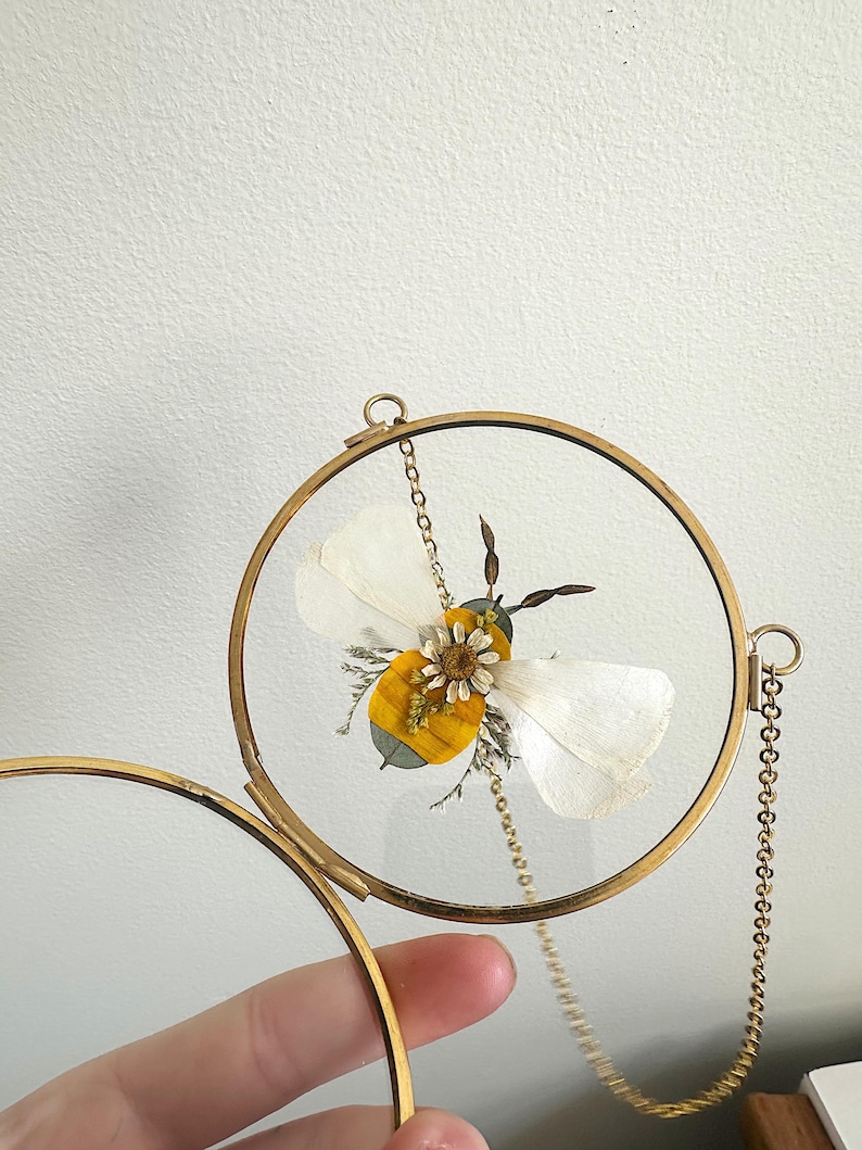 May include: A framed pressed flower art piece featuring a bee with white wings and a yellow body. The bee is encased in a round, gold-colored metal frame with a glass front, suspended by a delicate chain.