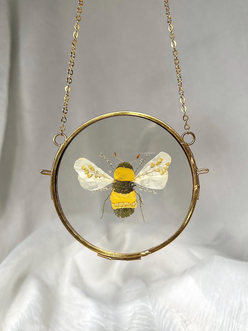 May include: A circular brass frame hanging from a gold-toned chain, enclosing a pressed flower art piece. The artwork features a bee created from dried yellow and green botanical elements, with white petals forming the wings, set between two glass panes.