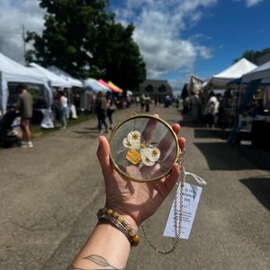 May include: A circular brass frame containing pressed white flowers and yellow wood pieces, suspended by a gold-toned chain. A white rectangular tag is attached to the chain, featuring printed text that reads: HI, I'M A BOTANICAL BUG! Bee!