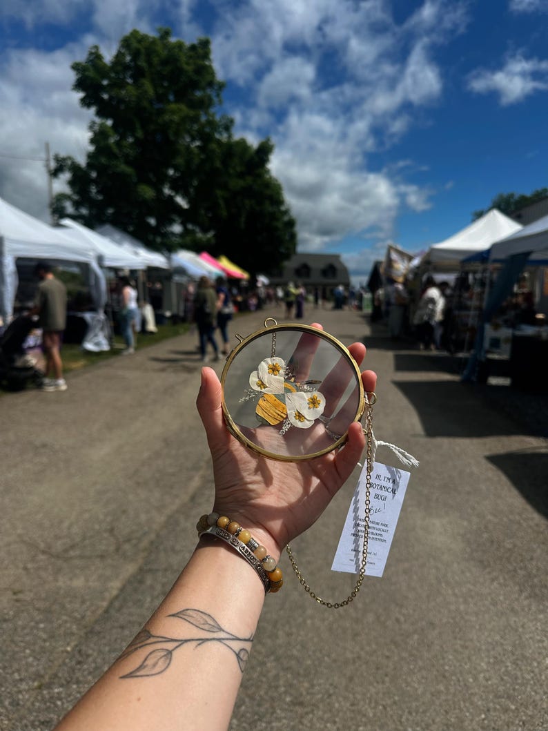 May include: A circular brass frame containing pressed white flowers and yellow wood pieces, suspended by a gold-toned chain. A white rectangular tag is attached to the chain, featuring printed text that reads: HI, I'M A BOTANICAL BUG! Bee!