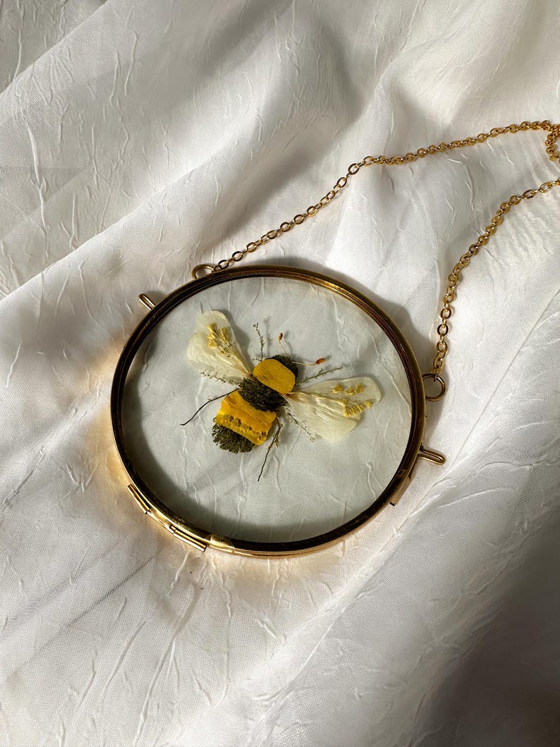 May include: A circular wall hanging featuring a pressed bee made of yellow and green dried botanical elements. The bee is encased between two clear glass panes within a thin gold-toned metal frame, suspended by a matching gold-toned metal chain.
