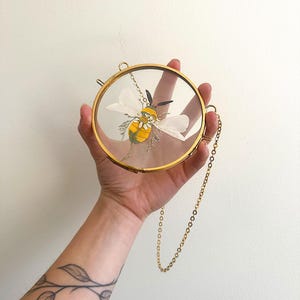 May include: A circular brass frame containing a pressed yellow and black bee illustration with white wings and small floral accents. The frame features a thin gold-toned metal chain attached to the side for hanging, showcasing a clear glass display.