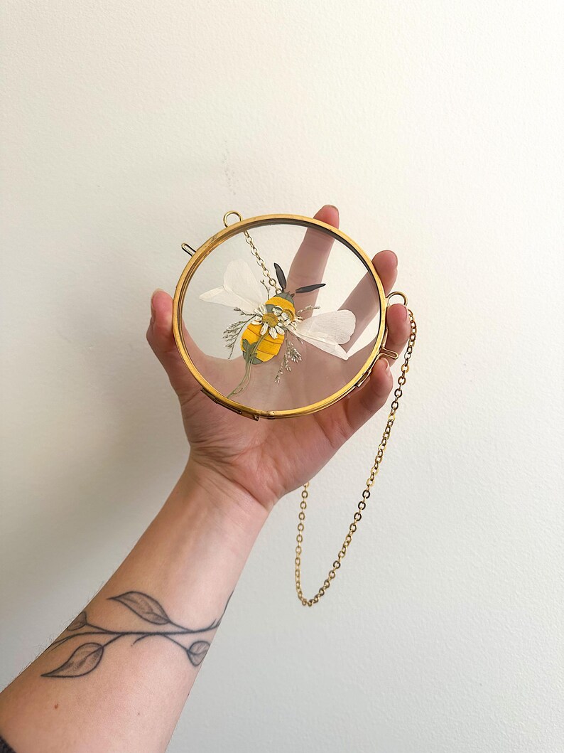 May include: A circular brass frame containing a pressed yellow and black bee illustration with white wings and small floral accents. The frame features a thin gold-toned metal chain attached to the side for hanging, showcasing a clear glass display.