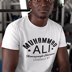 Puede incluir: Una camiseta blanca con un estampado gráfico negro que dice "Muhammad Ali Campeón de peso pesado Louisville KY".