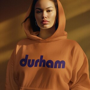 Pu&ograve; includere: Una persona indossa una felpa con cappuccio color arancio bruciato con la parola "durham" stampata in viola sul davanti.