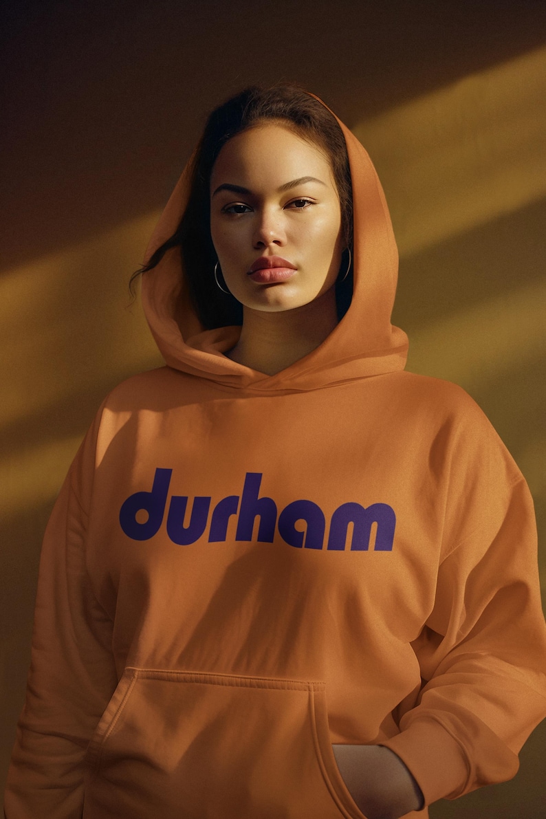 Pu&ograve; includere: Una persona indossa una felpa con cappuccio color arancio bruciato con la parola "durham" stampata in viola sul davanti.