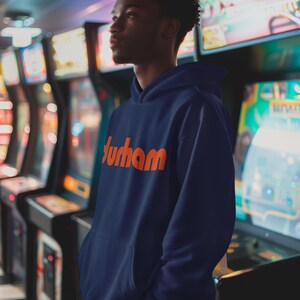 Pu&ograve; includere: Una persona indossa una felpa con cappuccio blu navy con la parola "Durham" stampata in arancione sul davanti. La persona &egrave; in piedi di fronte a una fila di giochi arcade.