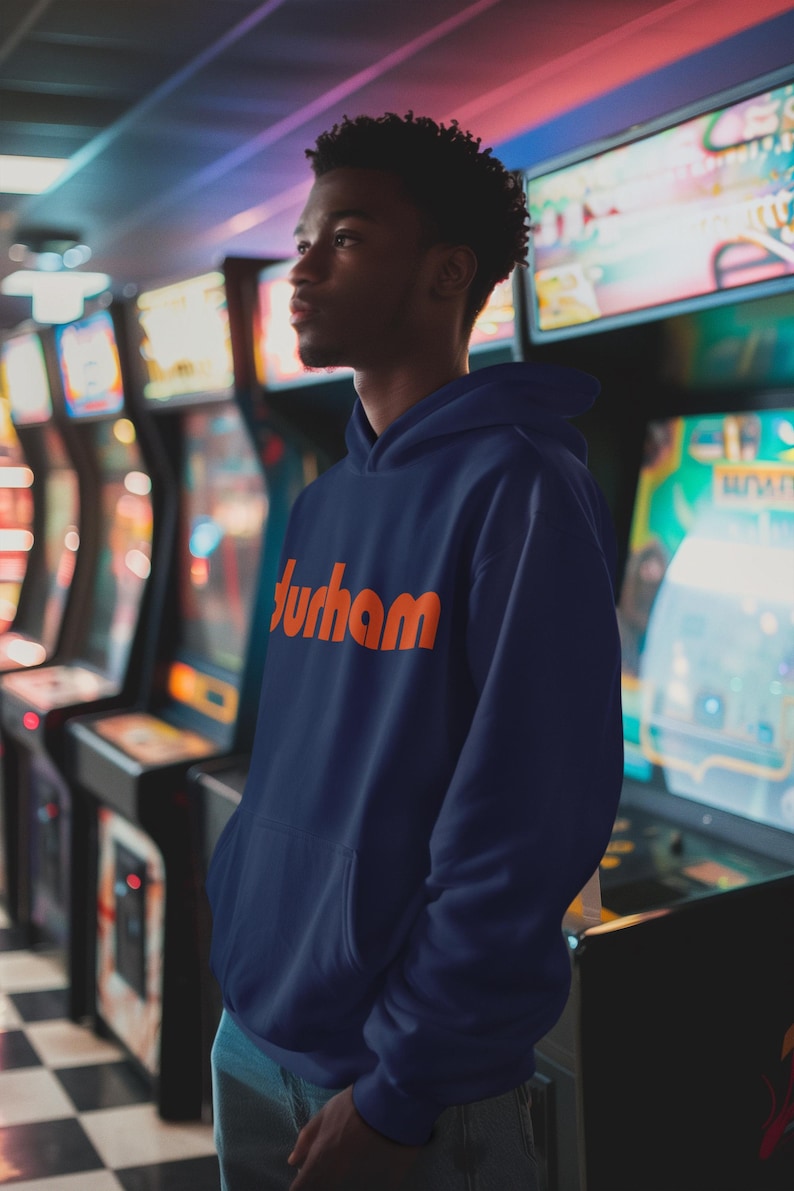 Pu&ograve; includere: Una persona indossa una felpa con cappuccio blu navy con la parola "Durham" stampata in arancione sul davanti. La persona &egrave; in piedi di fronte a una fila di giochi arcade.