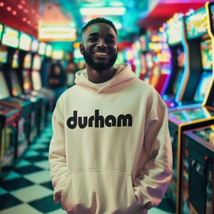 Pu&ograve; includere: Un uomo indossa una felpa con cappuccio bianca con la scritta "durham" stampata in nero sul davanti. &Egrave; in piedi in un'arcadia scarsamente illuminata con luci colorate sullo sfondo.