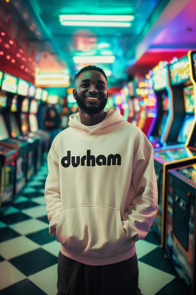 Pu&ograve; includere: Un uomo indossa una felpa con cappuccio bianca con la scritta "durham" stampata in nero sul davanti. &Egrave; in piedi in un'arcadia scarsamente illuminata con luci colorate sullo sfondo.