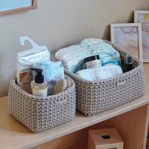 Peut inclure: Deux paniers de rangement en crochet beige remplis d'articles de soins pour bébés. L'un contient des couches, des lingettes et des produits de soin. L'autre contient du savon pour bébé, une brosse et d'autres essentiels. Une illustration d'abeille encadrée est accrochée au mur.