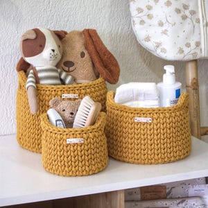 Peut inclure: Des paniers en crochet jaune moutarde de différentes tailles, remplis d'articles pour bébés et de peluches. Les paniers sont posés sur une surface blanche, avec un mur décoratif et un article à motifs floraux en arrière-plan. Les paniers portent de petites étiquettes "LITTLE MOSS".