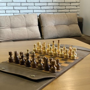 Könnte beinhalten: Ein braunes, ledergebundenes Schachbrett mit einem Schachspiel aus Holz. Die Schachfiguren sind in der Ausgangsposition auf dem Brett angeordnet. Das Schachbrett ist geöffnet und liegt auf einem Holztisch.