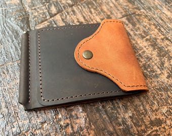 May include: A brown leather wallet with a tan leather flap and a metal button. The wallet has a money clip on the side.