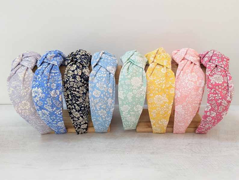 May include: A set of ten knotted headbands in various colors with a floral pattern. The headbands are displayed on a wooden stand.
