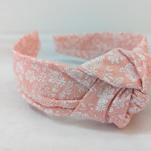 May include: A pink headband with a white floral pattern. The headband is tied in a knot at the top.