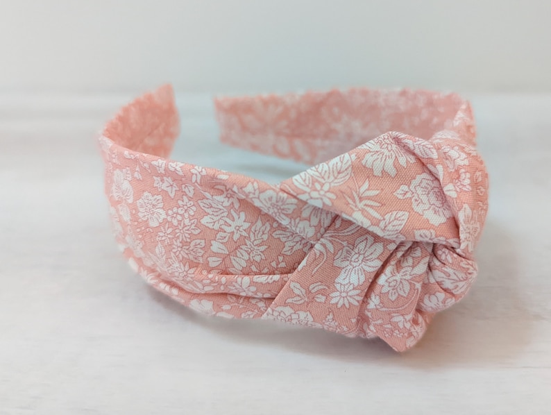 May include: A pink headband with a white floral pattern. The headband is tied in a knot at the top.